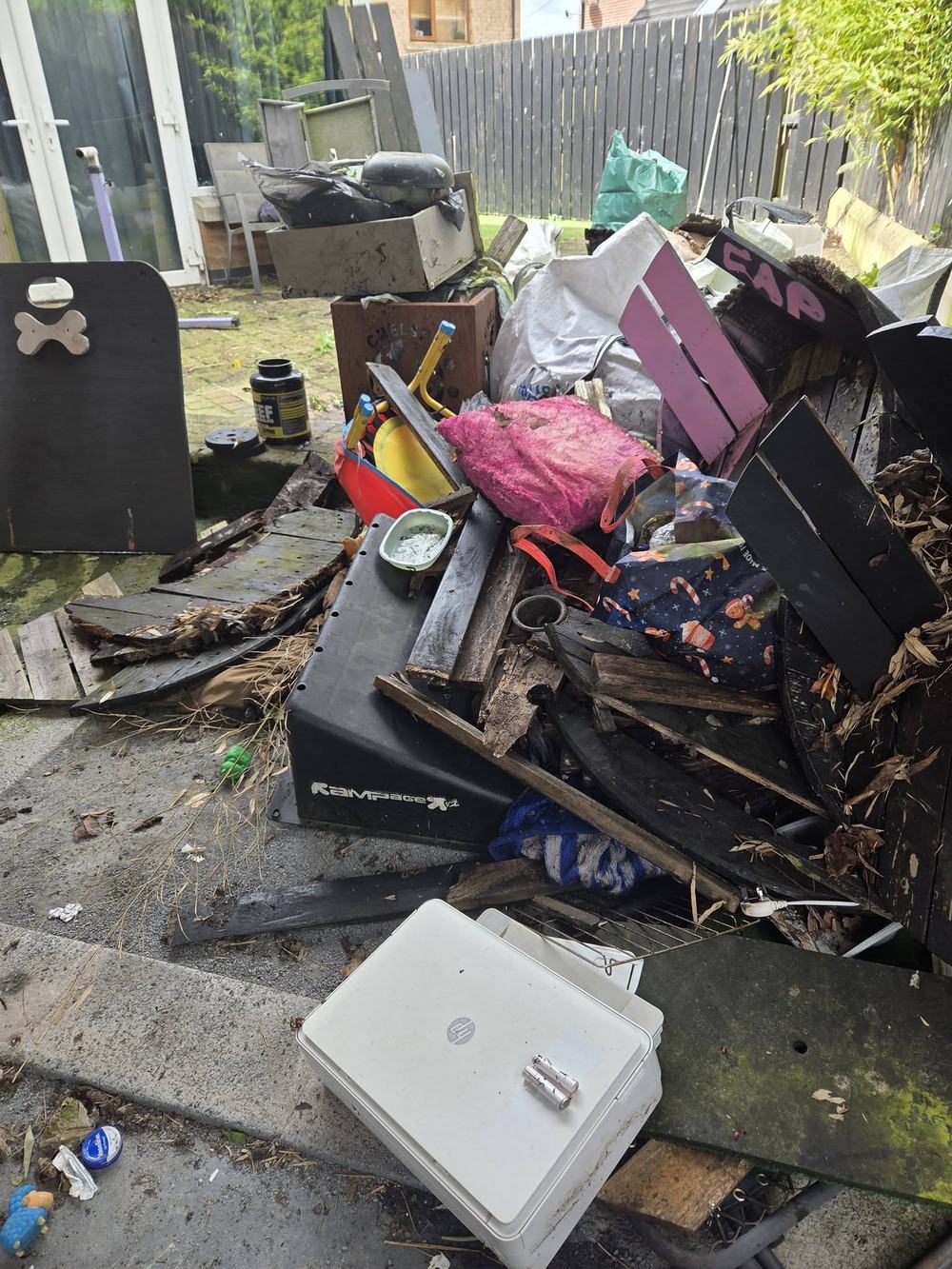 A back garden patio piled with broken decking, pallets, bags of waste and old furniture.